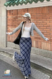 Woman in a white top and blue plaid pants standing on steps with a brick wall and green roof tiles in the background.