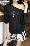 A woman wearing a black sheer off-the-shoulder long-sleeve top and black-and-white striped shorts, accessorized with a silver cross necklace, standing in a shop setting.