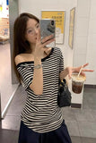 A woman takes a mirror selfie in a store while wearing a black and white horizontally striped off-the-shoulder t-shirt and a dark navy skirt. She is holding an iced coffee and wearing a black quilted shoulder bag.
