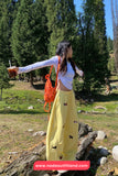 Woman in yellow pants and white top with orange bag in a forest setting, holding a drink.
