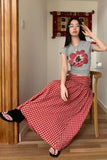 Woman wearing a gray t-shirt with a red flower design and a red checkered skirt, sitting on a wooden stool.