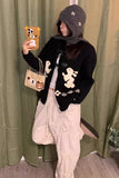 Person wearing a black hoodie with bear designs, holding a phone with a bear case, standing in front of a green curtain.
