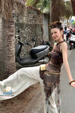 Woman in a patterned dress standing on a street with palm trees and scooters in the background.
