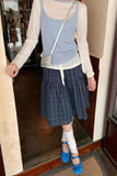 Person wearing a light gray long-sleeve shirt, blue plaid skirt, and blue shoes in a room.