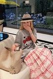 Woman sitting on a couch with a straw bag, wearing a hat and a gray sweater with a red bow design.