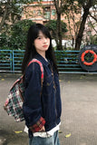 A young woman standing outdoors in front of trees and a blue railing, wearing a dark blue denim jacket with red plaid cuffs, a red, green, and white plaid backpack, and light blue jeans.