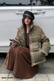 Person wearing a plaid coat and brown skirt sitting next to a white vehicle.