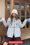 Woman wearing a patterned cardigan and white beanie in front of a wooden building.