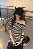 A woman wearing a black and white horizontally striped off-the-shoulder t-shirt, a black bucket hat, and a black quilted handbag with a white logo, standing in an indoor modern space.