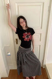 Person wearing a black t-shirt with a red flower design and a checkered skirt standing against a white door.