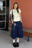 A woman stands outdoors wearing a pale yellow graphic T-shirt, dark blue polka-dot wide-leg cropped pants, white socks, and black loafers, with a black shoulder bag. The background features a textured terracotta wall and a wooden bench.