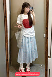 Woman taking a mirror selfie wearing a white t-shirt with a red pocket design, light blue floral skirt, and red sneakers.