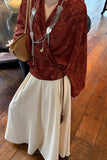 Person wearing a rust-colored top with intricate details and a beige skirt on a wooden floor.