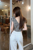 Woman in a checkered top and white pants standing in a modern interior setting.