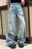 Light blue jeans with black designs worn by a person, standing against a wooden panel background.