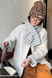 Person wearing a leopard print beanie, white coat, and striped scarf holding a handbag.