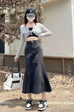 A woman stands outdoors wearing a gray long-sleeve knit crop top over a black bralette, paired with a high-waisted dark gray denim skirt. She is also wearing a black baseball cap, a white face mask, black and white sneakers, and holding a white tote bag.