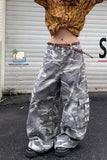 Person wearing camouflage pants with a plaid shirt, standing in front of a white wall with a logo.