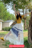 Woman in a yellow top and white skirt with blue patterns standing outdoors under trees.
