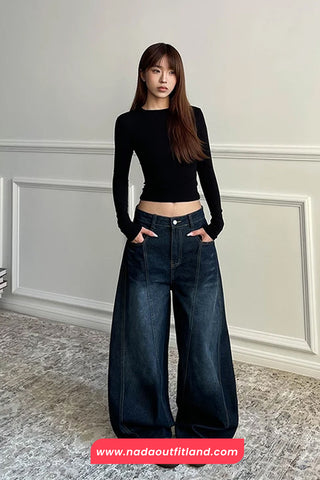 Person wearing a black long-sleeve top and wide-leg jeans standing against a light gray wall.