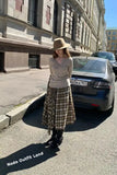 Woman in a beige sweater and plaid skirt standing on a street with a building in the background.