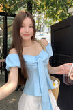 Woman in a light blue top and white pants holding a drink outdoors.