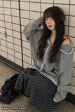 Woman sitting against a tiled wall wearing a gray sweater and plaid skirt, with headphones on.