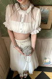 Person wearing a white outfit with a pleated skirt and sheer top, holding a decorative clutch.