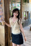 A woman wearing a cream-colored ribbed knit V-neck short-sleeve top and a dark denim mini skirt, holding an iced coffee in a rustic cafe setting.
