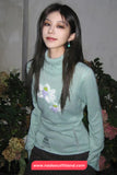 Woman wearing a light green sweater with floral embroidery, standing in front of flowers.