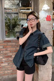 A young woman wearing a dark blue denim short-sleeve button-up shirt and black shorts, standing outdoors next to a brick wall and stone pillar. She is holding a black shoulder bag and wearing glasses.