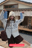 Woman wearing a white patterned cardigan and beige beanie outdoors with a wooden building in the background.