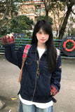 A young woman wearing a dark blue denim jacket with red plaid lining on the cuffs and collar, layered over a white t-shirt and paired with a brown shoulder bag, standing outdoors in front of trees and a railing.