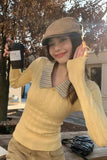 Person wearing a yellow sweater and beige cap, holding a coffee cup outdoors.