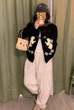 Person wearing a black sweater with bear designs, beige pants, and a beige bag, standing in front of a green curtain.