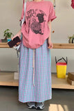 Person wearing a pink t-shirt with a graphic design and a long plaid skirt in an indoor setting.