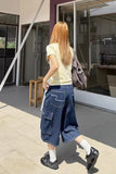A woman with long strawberry blonde hair is seen from behind, wearing a pale yellow t-shirt, a dark blue patterned cargo skirt, white socks, and black shoes, carrying a gray patterned shoulder bag while walking outdoors.