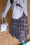 Person wearing a gray hoodie and plaid skirt with a small white bag on a wooden floor.
