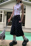 Person wearing a sleeveless floral top and polka dot skirt holding a small bag, standing in front of a house.
