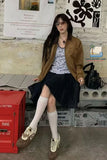 Woman in a brown coat and black skirt sitting on a bench with a red box and posters in the background.
