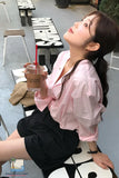 Woman in a pink shirt and black skirt sitting outdoors with a drink, wearing a gold earring.