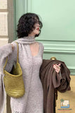 Woman in a gray outfit holding a yellow bag and a brown coat against a light green door.