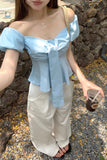 Person wearing a light blue off-shoulder top and beige pants, standing against a stone wall.