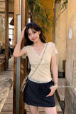 A woman posing in a rustic indoor setting, wearing a beige lace V-neck short-sleeve top, a dark denim mini skirt, and a blue headband.