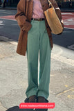 Person wearing a brown coat, pink shirt, and green pants on a street.