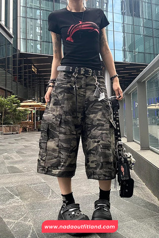 Person wearing a black t-shirt with a red graphic, camouflage cargo shorts, and black Crocs in an urban setting.