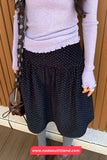 Person wearing a light purple sweater and dark polka dot pants, holding a brown bag.