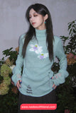 Woman wearing a light blue sweater with floral embroidery in front of plants.