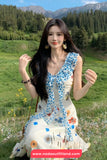 Woman in a floral dress sitting in a scenic outdoor setting with mountains and trees.