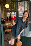 A woman posing in a cafe setting wearing a dark blue denim short-sleeve button-up shirt, a brown pleated mini skirt, brown lace-up boots, and carrying a small brown handbag.